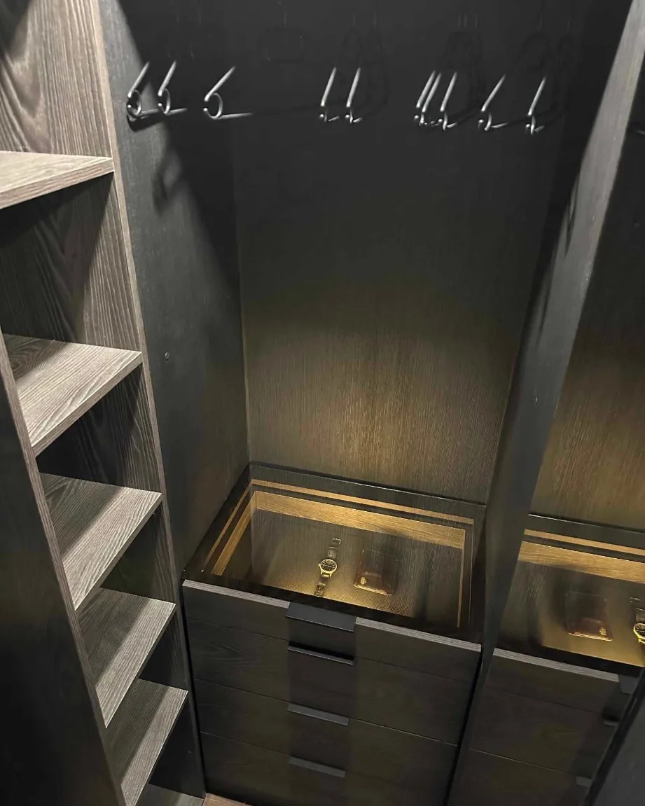 Wnętrze ciemnej, drewnianej garderoby z półkami i szufladami. W szufladzie podświetlonej od góry leży zegarek i portfel.