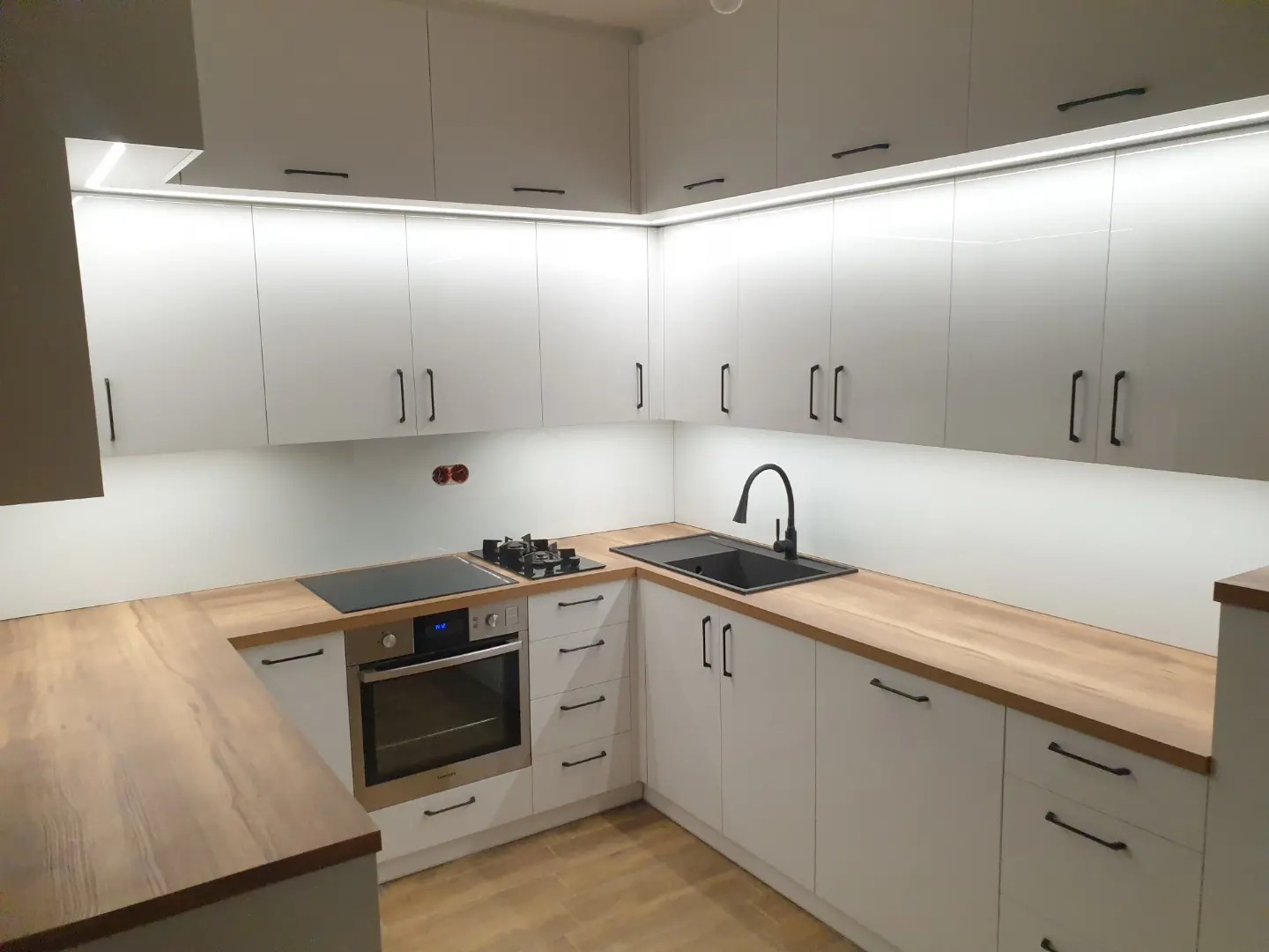 Nowoczesna kuchnia z białymi szafkami, drewnianym blatem i czarnymi uchwytami. Podświetlenie LED pod szafkami górnymi. Na blacie płyta indukcyjna i zlewozmywak z czarną baterią.