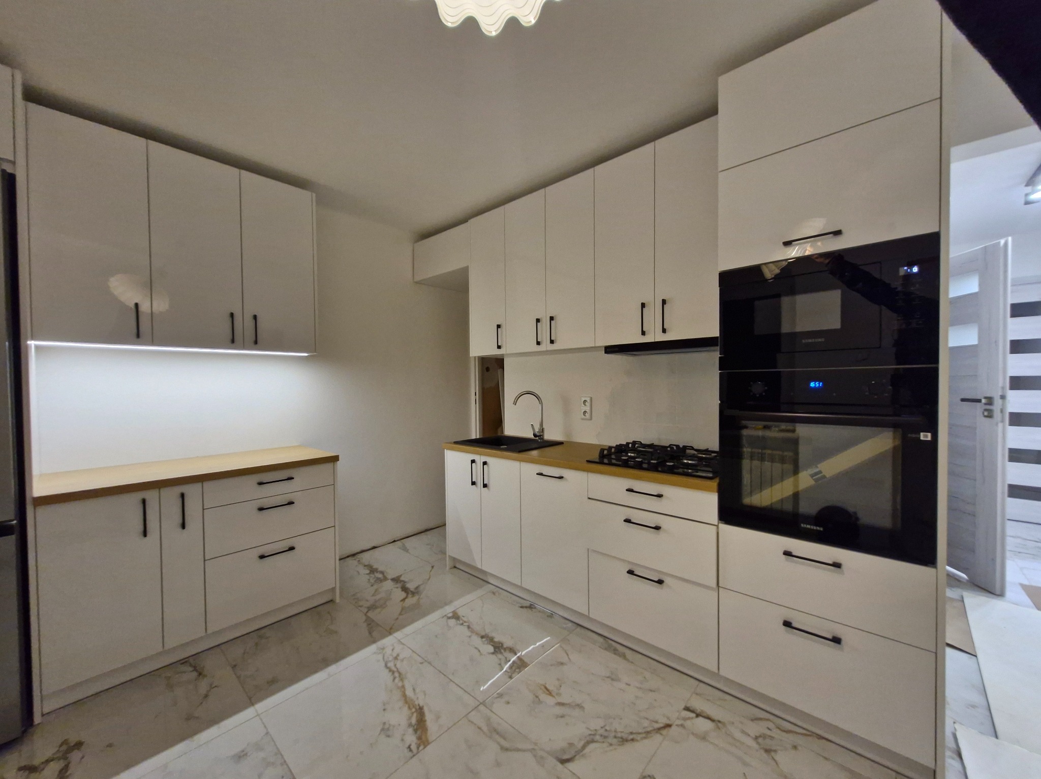Nowoczesna kuchnia z białymi szafkami i drewnianym blatem, czarnymi uchwytami, marmurową podłogą i wbudowanym piekarnikiem Samsung. Oświetlenie LED pod szafkami.