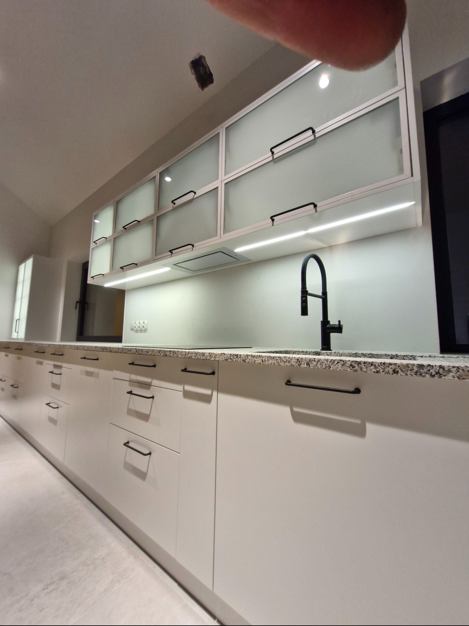 Nowoczesna, minimalistyczna kuchnia z białymi frontami, czarnymi uchwytami i blatem z jasnego granitu. Górne szafki z matowego szkła, oświetlenie LED pod szafkami.