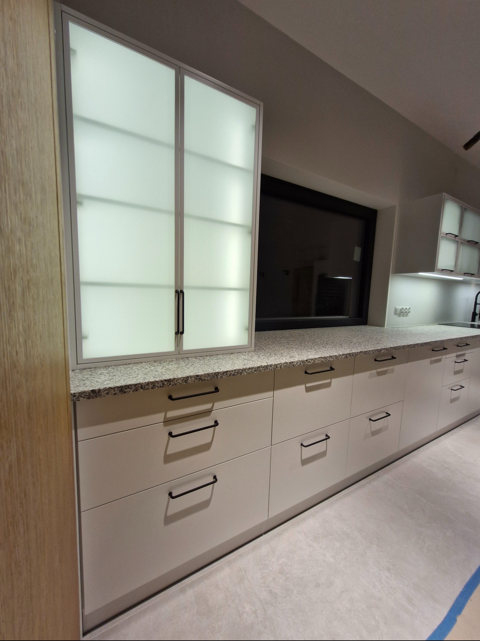 Nowoczesna kuchnia z białymi szafkami i blatem z granitu, czarne uchwyty, górne szafki z mlecznym szkłem, oświetlenie LED pod szafkami, minimalistyczny design.