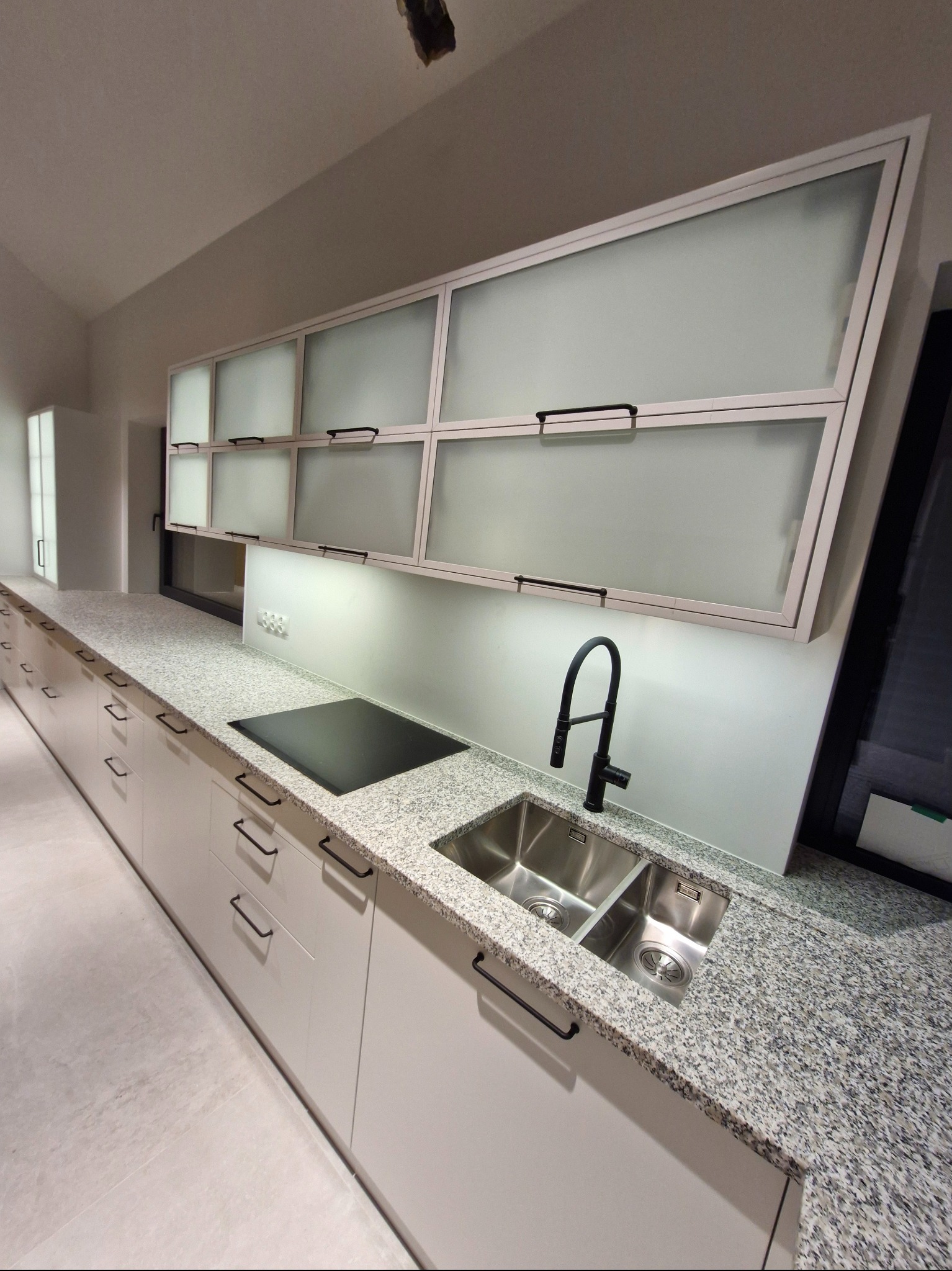 Nowoczesna kuchnia z białymi szafkami, blatem z szarego granitu i czarną armaturą. Górne szafki z mlecznego szkła podświetlone LED. Minimalistyczny design.