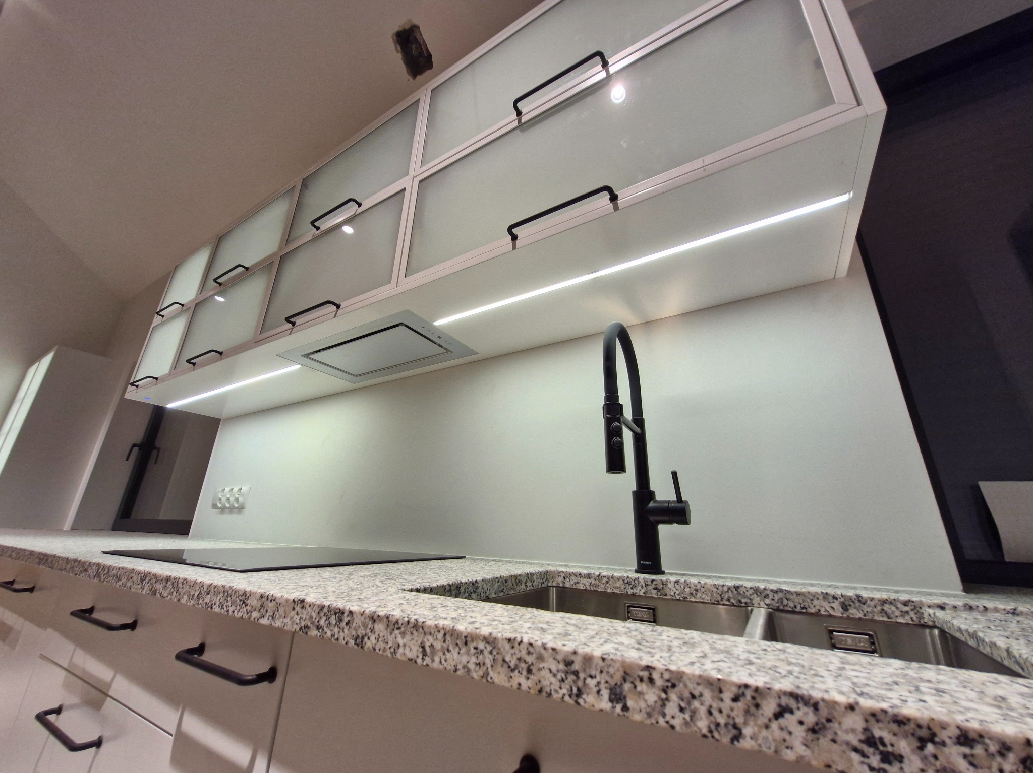 Nowoczesna kuchnia z białymi szafkami z matowego szkła, czarnymi uchwytami i granitowym blatem. Oświetlenie LED podkreśla minimalistyczny design i funkcjonalność.