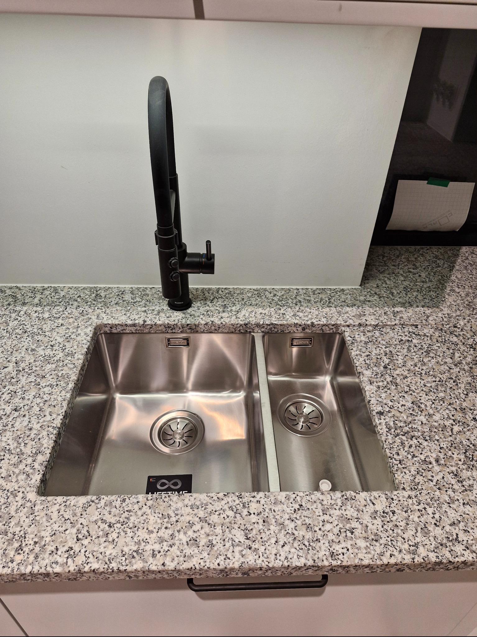 Dwukomorowy, stalowy zlew z czarną baterią na blacie z jasnego granitu. Widoczne logo producenta na dnie zlewu. Nowoczesny design.