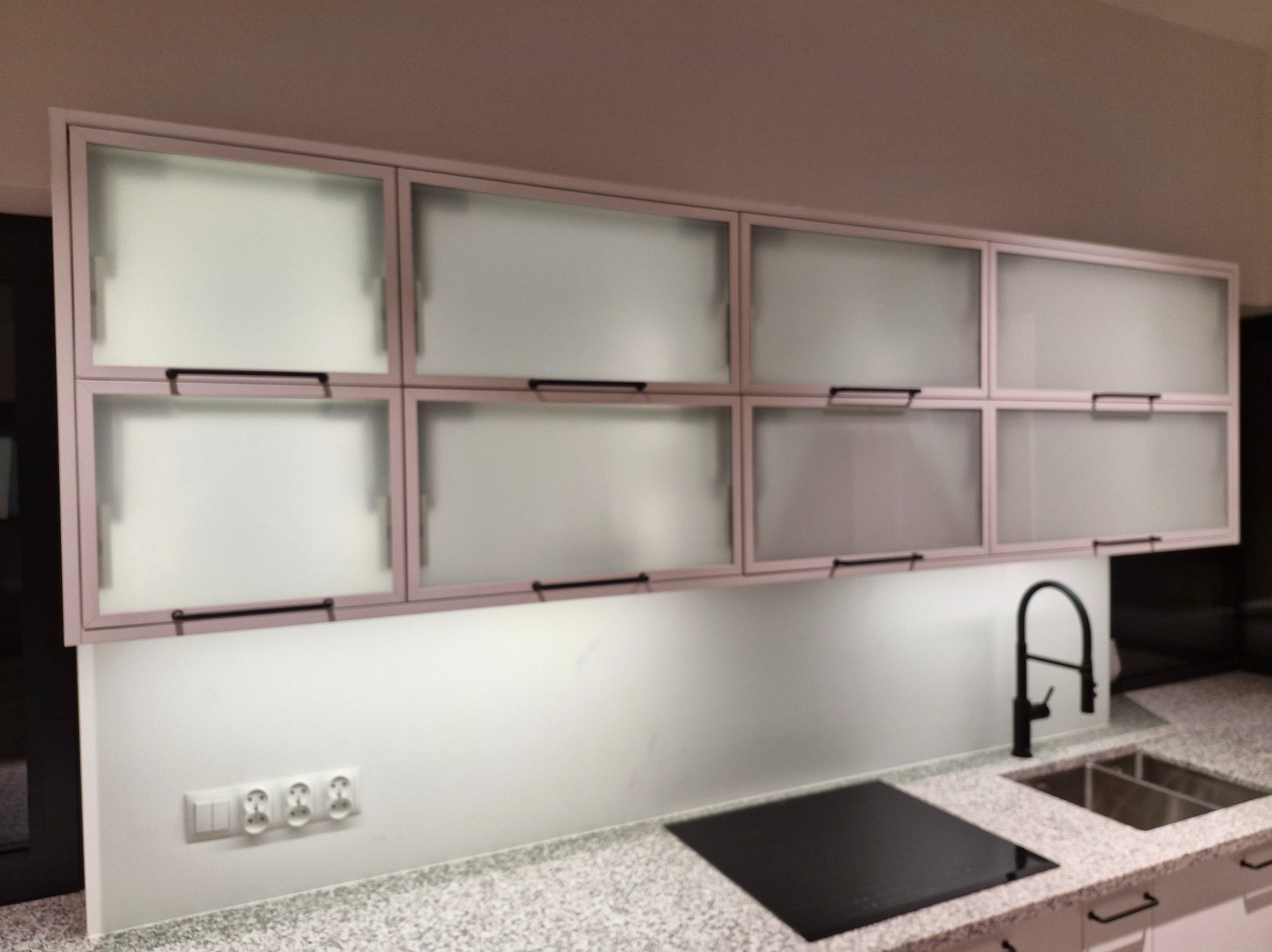 Nowoczesna kuchnia z górnymi szafkami z matowego szkła i czarnymi uchwytami, oświetleniem LED pod szafkami i granitowym blatem z czarną płytą indukcyjną.