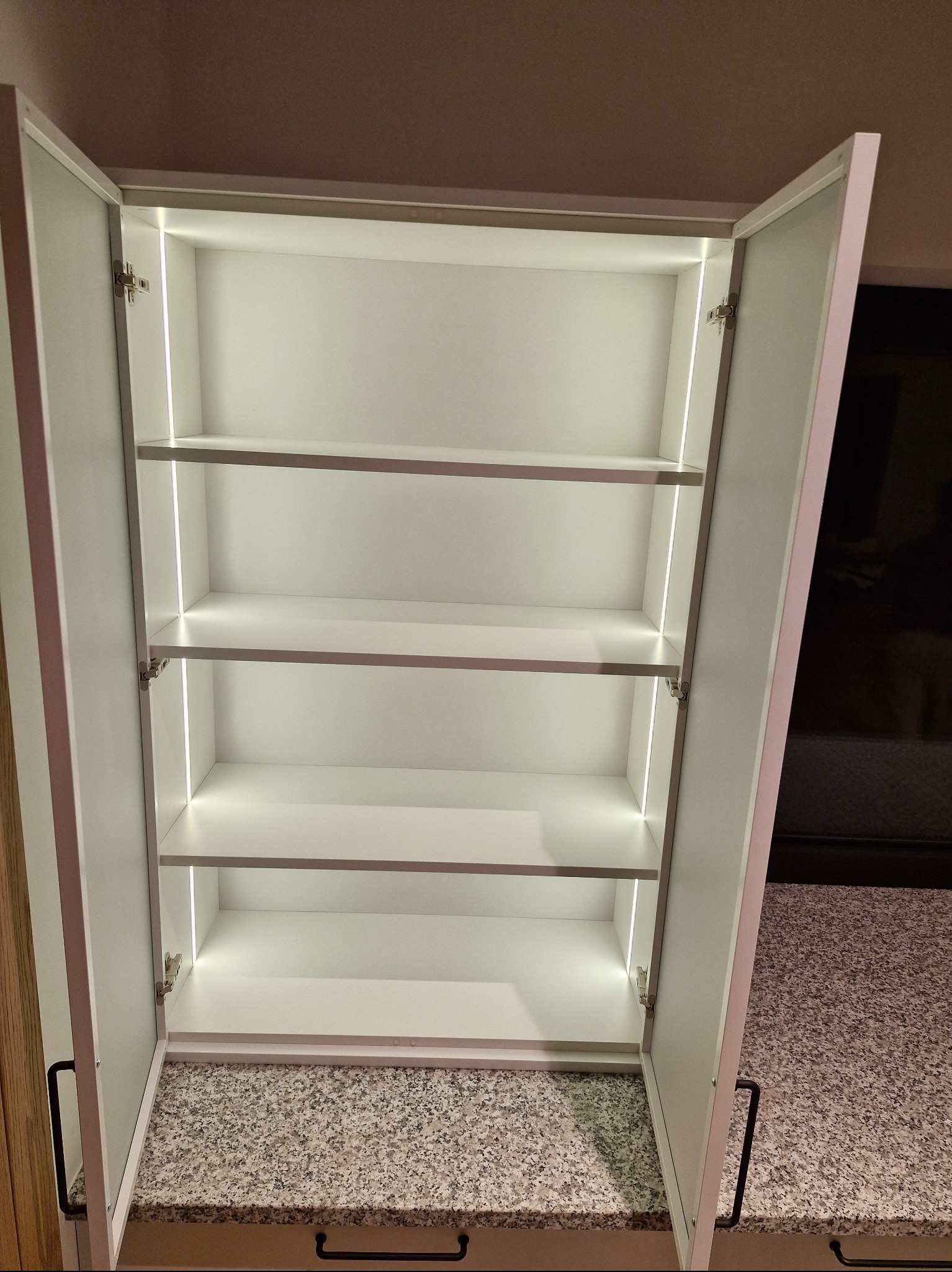 Otwarta, biała szafka z czterema półkami i subtelnym oświetleniem LED wzdłuż krawędzi, ukazująca wnętrze na tle blatu z szarego granitu.