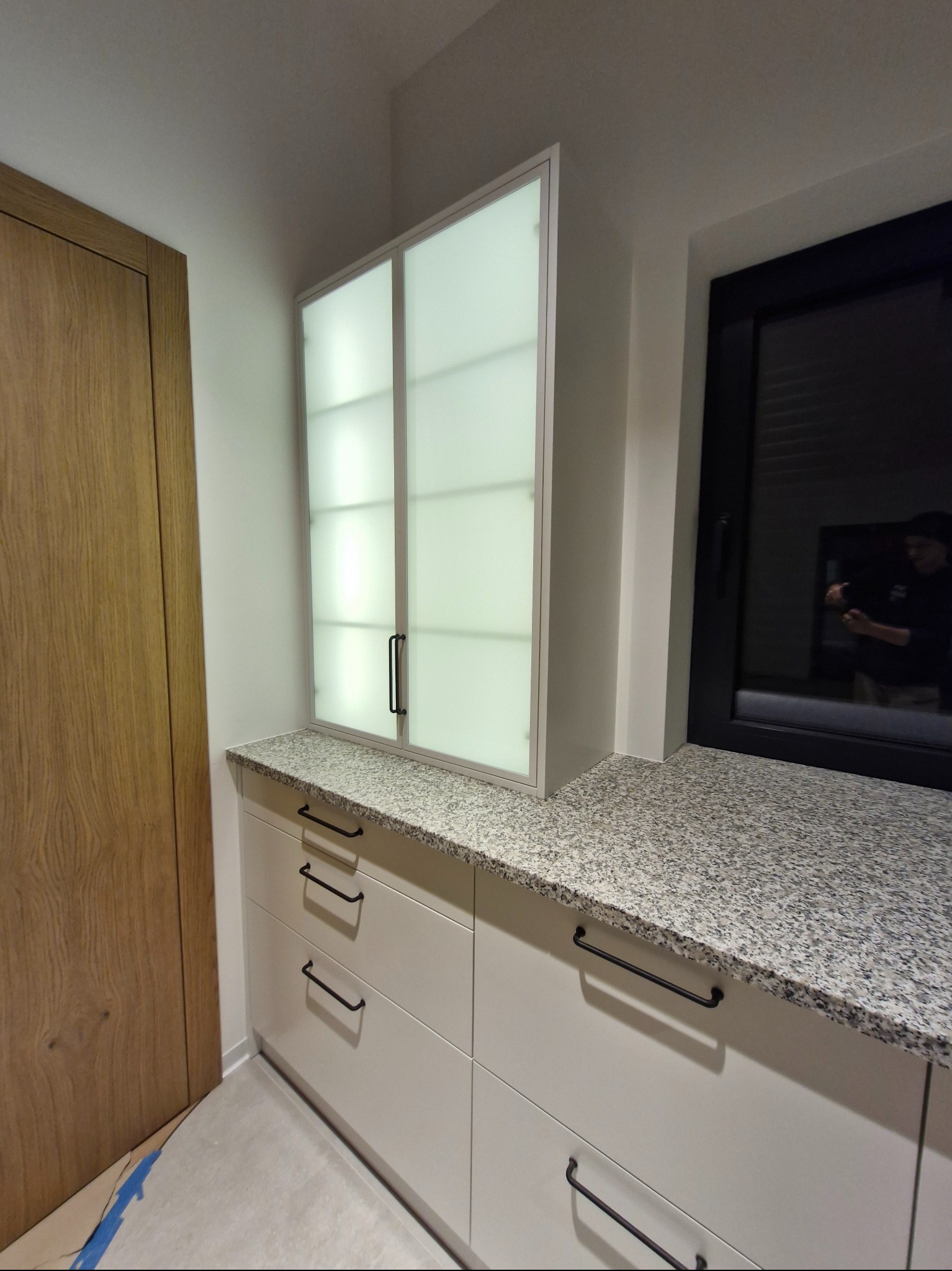 Jasna zabudowa kuchenna z szafkami z mlecznego szkła i blatem z szarego granitu, czarne uchwyty, widok na okno z odbiciem osoby robiącej zdjęcie.