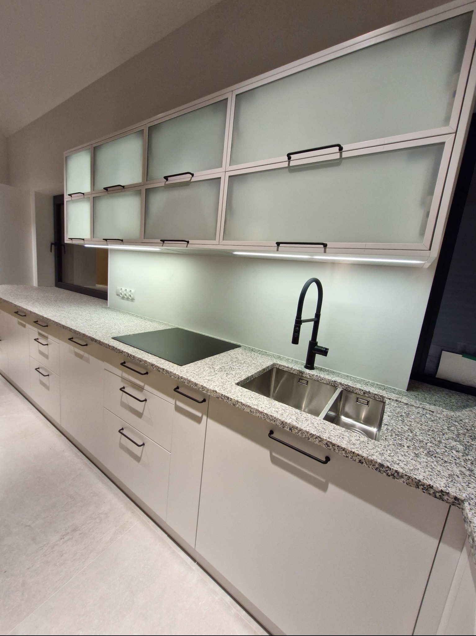 Nowoczesna kuchnia z białymi szafkami, blatem w plamki i czarną armaturą. Górne szafki z mlecznym szkłem i podświetleniem LED. Minimalistyczny design.