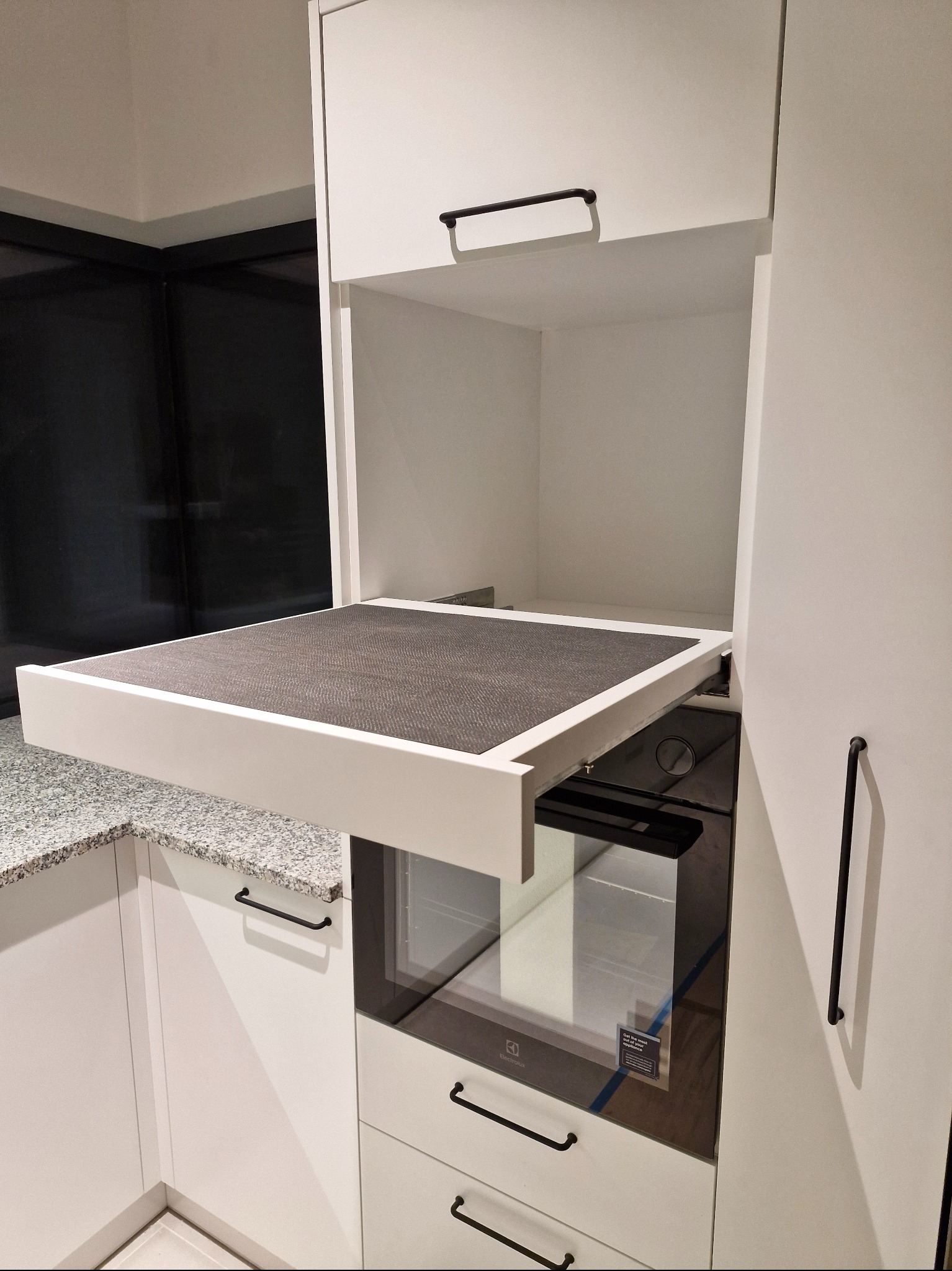 Biała kuchnia na wymiar z wysuwanym blatem roboczym nad piekarnikiem, czarne uchwyty, szary blat, nowoczesny design.