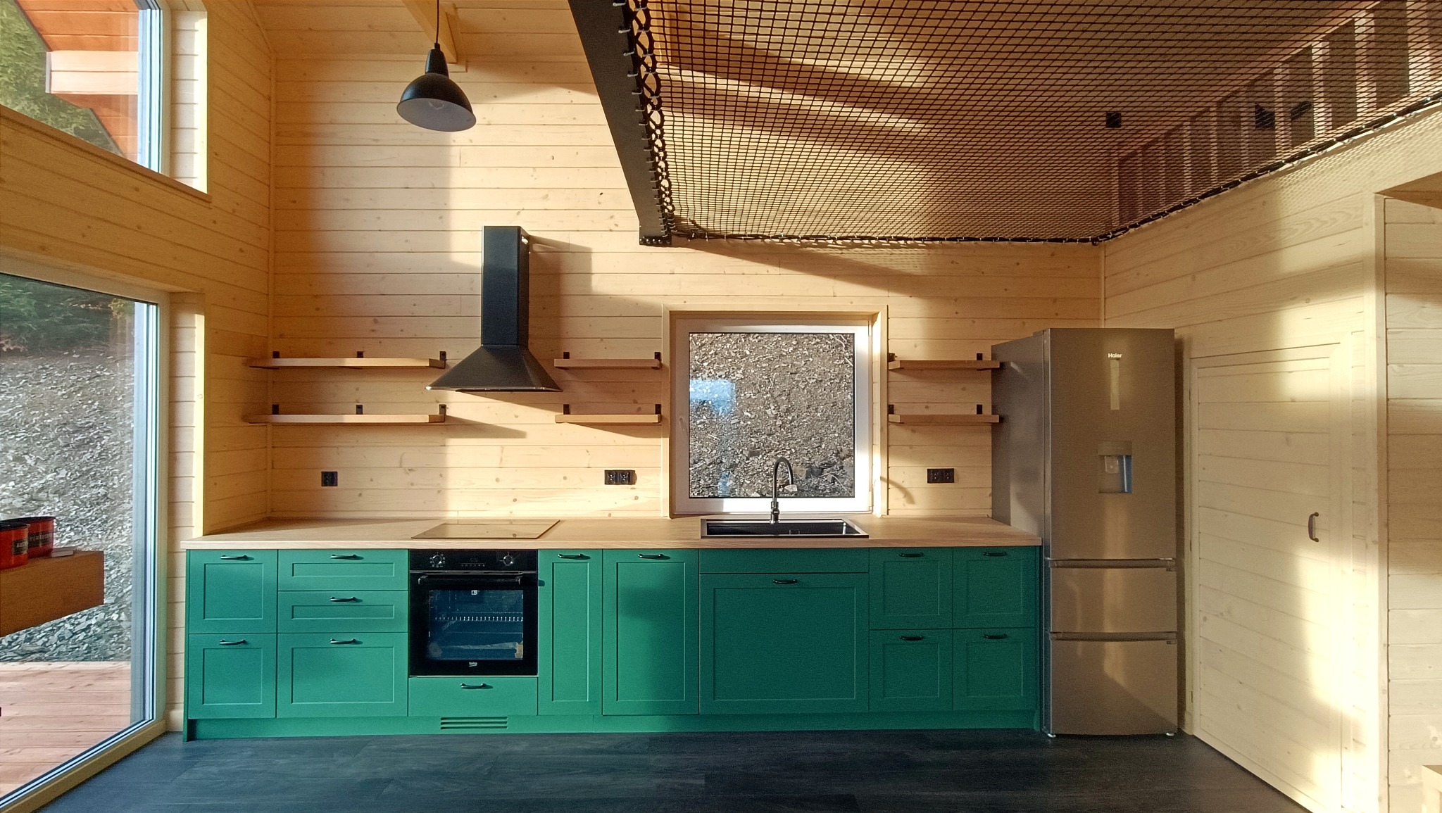 Nowoczesna kuchnia z zielonymi szafkami i drewnianymi ścianami. Nad oknem i lodówką siatka zabezpieczająca. Minimalistyczny design z naturalnymi materiałami.