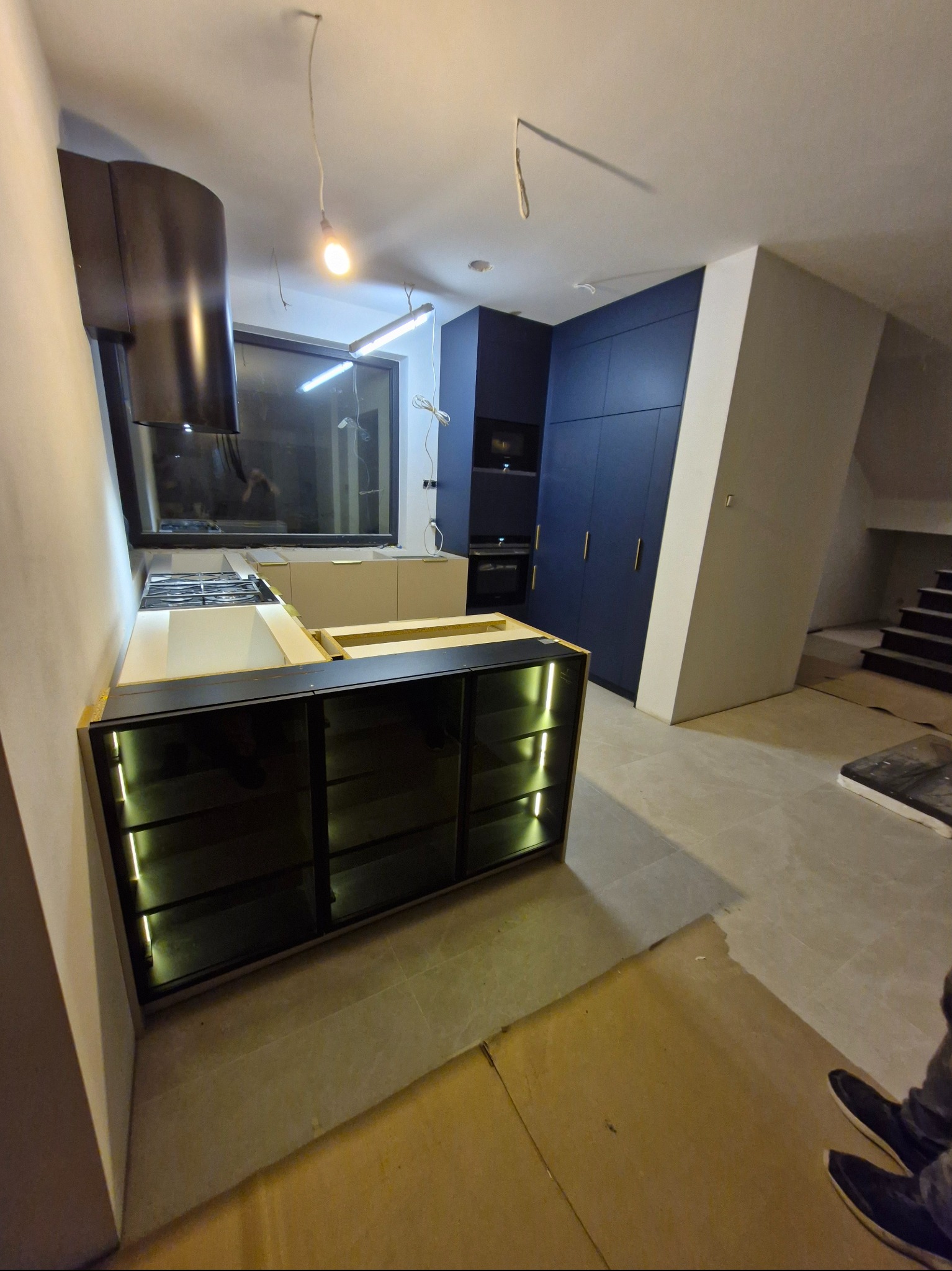 Nowoczesna kuchnia w trakcie montażu: czarne szafki z podświetleniem LED, białe blaty, ciemne szafy w zabudowie i okap. Widoczne elementy instalacji elektrycznej.