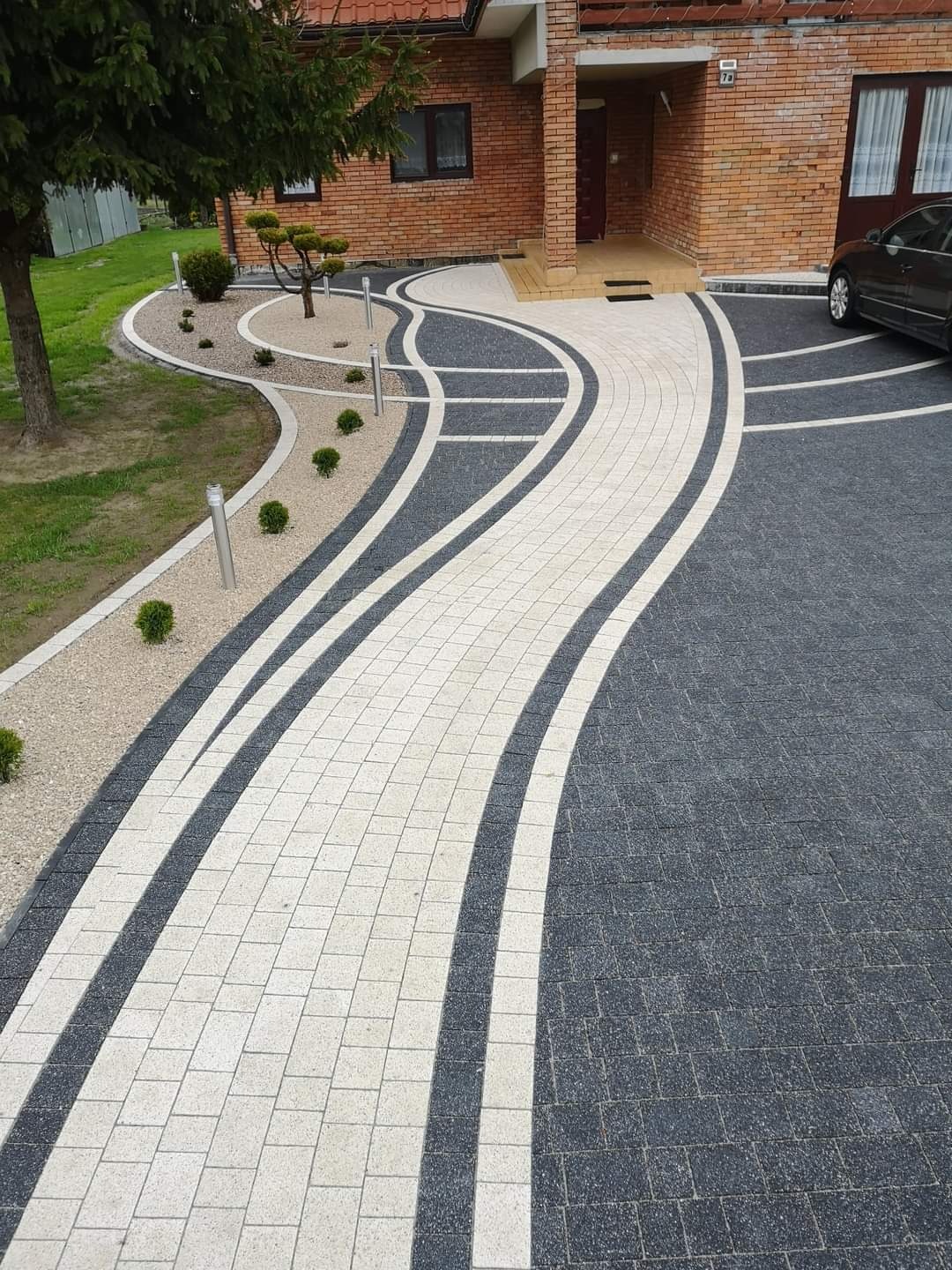 Zakrzywiony chodnik z kostki brukowej w odcieniach szarości i bieli prowadzący do domu z cegły, z elementami dekoracyjnymi z kruszywa i niskich krzewów.