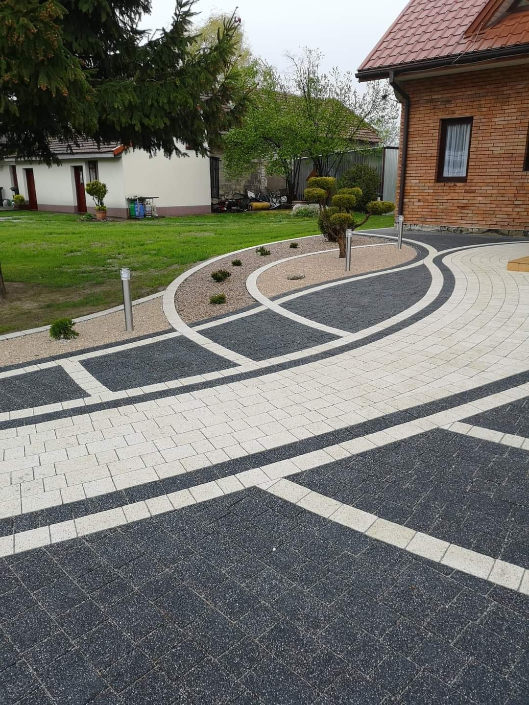 Ogród z nowocześnie ułożoną ścieżką z kostki w kolorach szarości i bieli, z elementami dekoracyjnymi z kamienia i roślin, oświetloną niskimi lampami, na tle domu z czerwonej cegły.