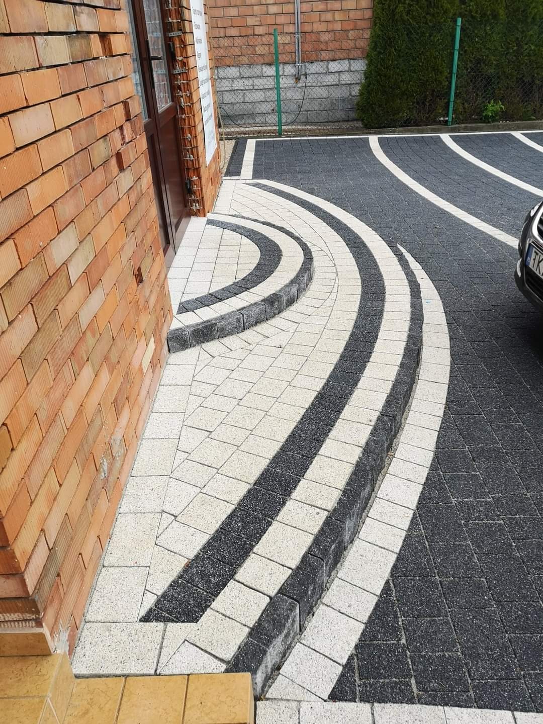 Ułożona nawierzchnia z kostki brukowej w kolorach czerni i bieli, tworząca łukowaty wzór przy budynku z cegły, z widocznym fragmentem zaparkowanego samochodu.