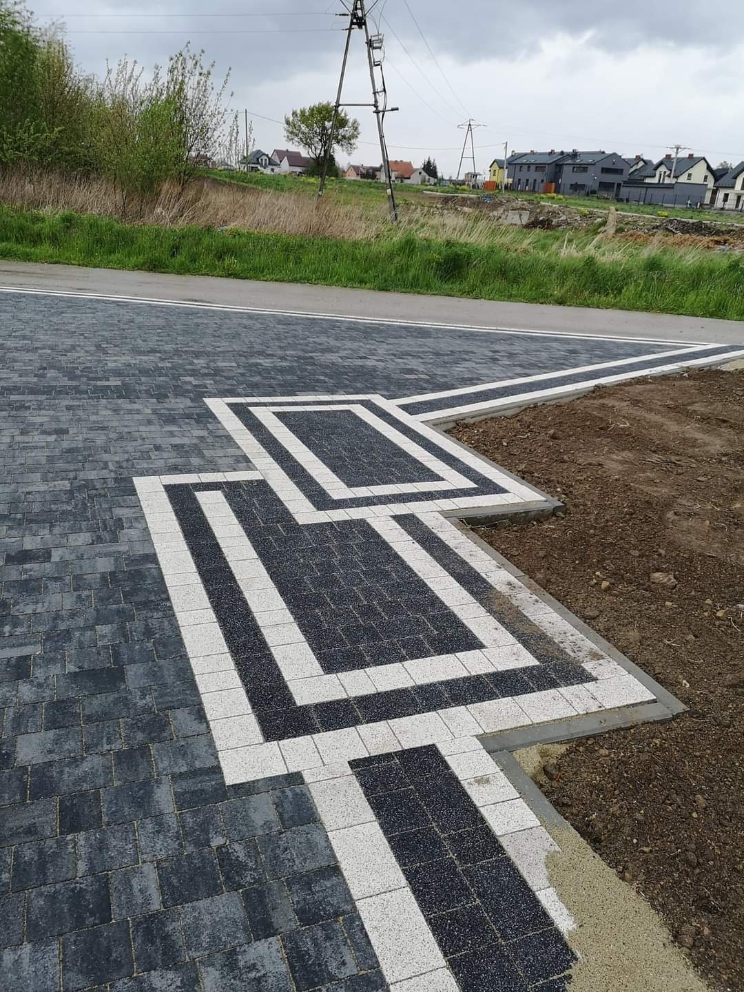 Ukończony chodnik z kostki brukowej w odcieniach szarości i bieli, ułożony we wzór z prostokątami, przylegający do asfaltowej drogi i ziemi, na tle zabudowań mieszkalnych i słupa wysokiego napięcia.
