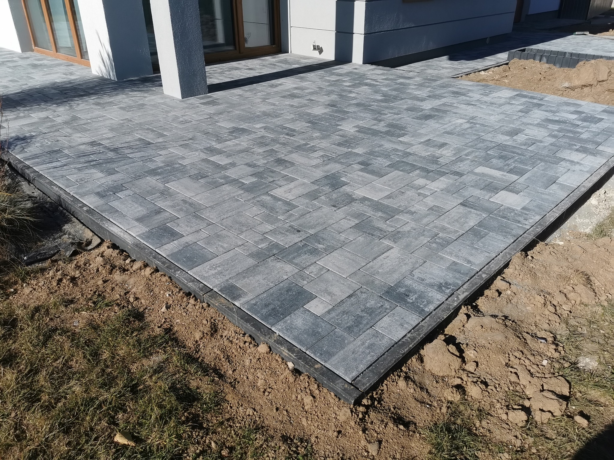 Ułożona szara kostka brukowa na posesji, widoczna krawędź z obrzeżem i przygotowane podłoże z ziemią.