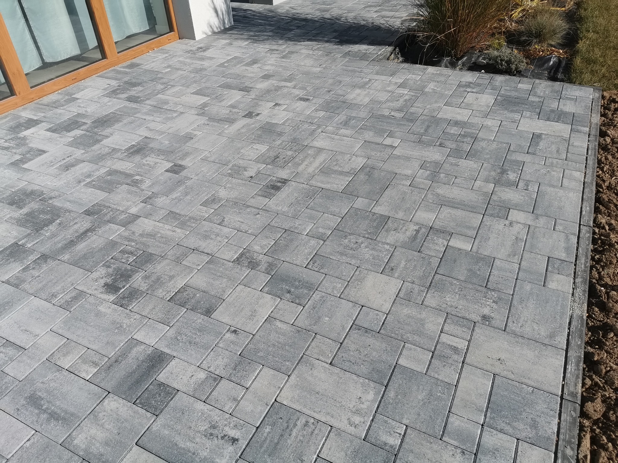 Nowoczesny, szary chodnik z kostki brukowej ułożonej w nieregularny wzór, przylegający do budynku z drewnianymi oknami i elementami roślinności.