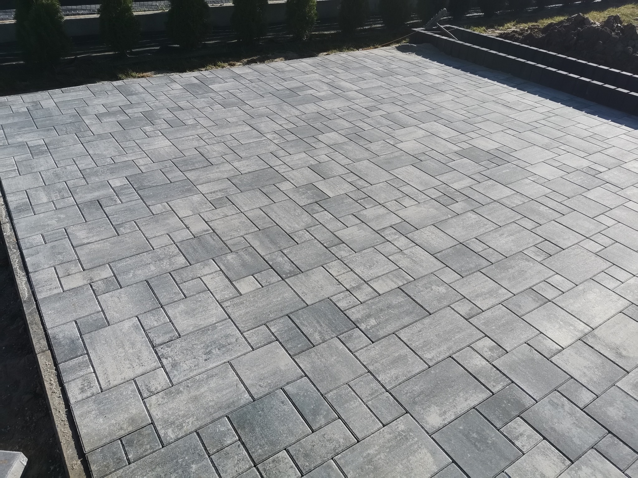 Ułożona szara kostka brukowa w geometryczny wzór, widoczne obrzeża trawnika i rząd tuj w tle.