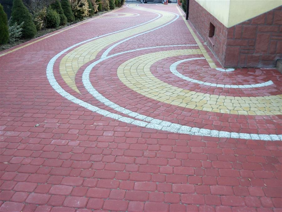 Wzorzysty podjazd z czerwonej kostki brukowej z dekoracyjnymi wstawkami z kostki żółtej i białej, tworzącymi faliste linie i okręgi przy budynku.