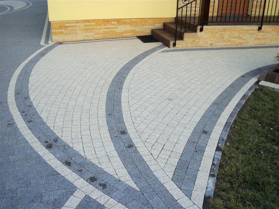 Ułożona z jasnoszarej i ciemnoszarej kostki brukowej ścieżka w kształcie łuku prowadząca do schodów wejściowych domu z żółtą elewacją i czarną balustradą.