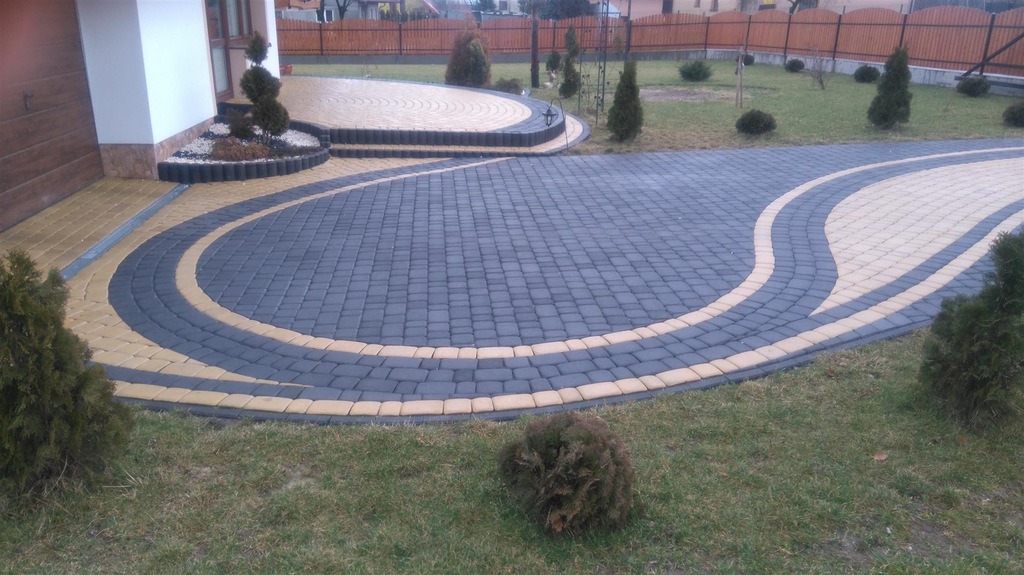 Ułożona nawierzchnia z kostki brukowej w kolorach szarym i beżowym, tworząca łukowate wzory na podjeździe, z widoczną roślinnością ozdobną i ogrodzeniem w tle.