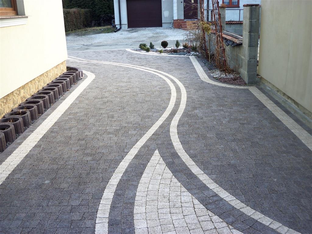 Szeroki podjazd z szarej kostki brukowej z dekoracyjnymi wstawkami z jasnej kostki, obrzeża z betonowych donic i jasnej kostki, widok z góry.