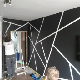 Pracownik w koszulce z logo LOTOS nakleja taśmę malarską na czarną ścianę z geometrycznym wzorem z białych linii, drabina i folia ochronna na podłodze, widok z perspektywy dziecka.