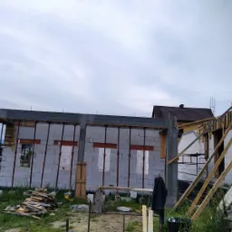 Wznoszenie ścian z betonu komórkowego na budowie domu jednorodzinnego, widoczne podpory szalunkowe i drewniane elementy konstrukcyjne dachu, w tle zabudowa mieszkalna.