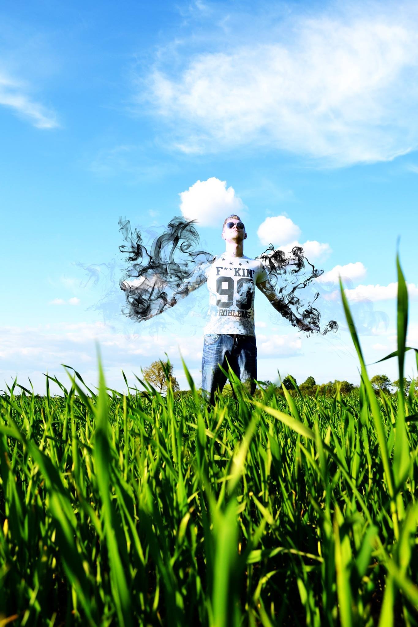 Mężczyzna w okularach przeciwsłonecznych stoi na polu zielonej trawy z rozpostartymi ramionami, z efektami dymu wokół ramion, na tle błękitnego nieba z chmurami.