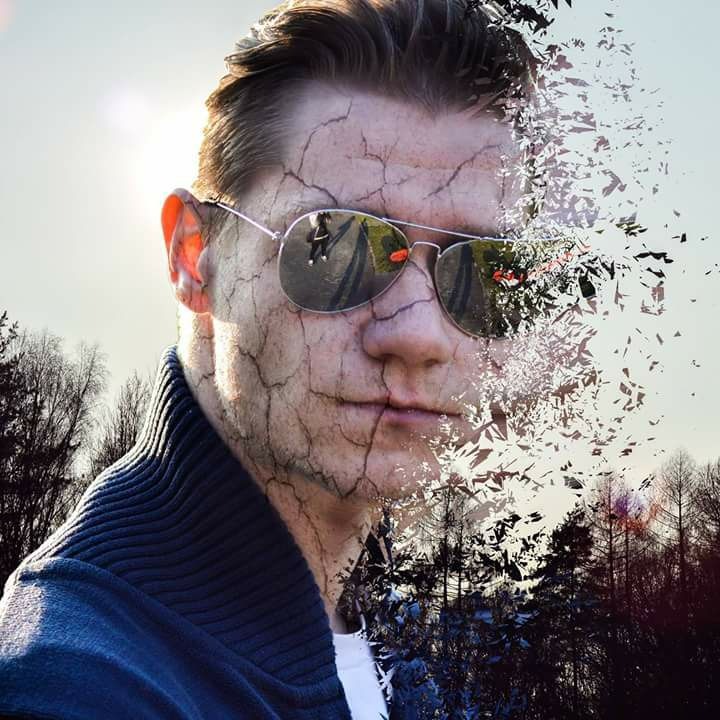 Portret mężczyzny w okularach przeciwsłonecznych z efektem pęknięć na skórze i rozpadu na drobne elementy, na tle drzew i nieba.