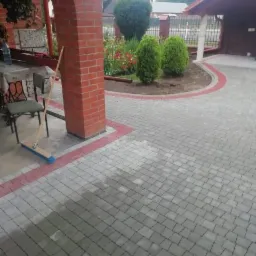 Ułożona szara kostka brukowa z czerwoną obwódką, widoczny fragment domu z ceglanym łukiem, krzesło, miotła oparta o filar i zadbany ogródek z tujami.