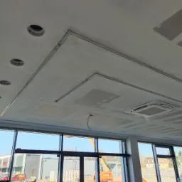 Sufit z widocznymi elementami konstrukcyjnymi pod płyty karton gips, otwory na oświetlenie i klimatyzację, widok przez okno na budynek i podnośnik koszowy.