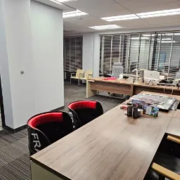 Wnętrze biura z biurkami, krzesłami i oknami z żaluzjami, z otwartymi drzwiami prowadzącymi do schodów, gdzie widoczne są materiały budowlane.