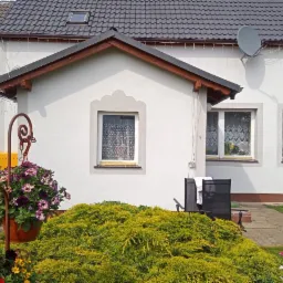 Dom jednorodzinny z białą elewacją, szarą dachówką i dekoracyjnymi obramowaniami okien, widok z ogrodu z kwitnącymi kwiatami i ozdobną fontanną.