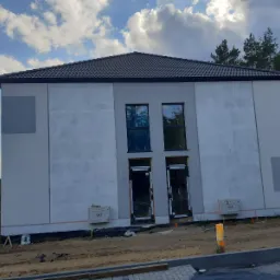 Nowoczesny dom jednorodzinny w trakcie ocieplania elewacji, widoczne fragmenty nieotynkowanej powierzchni i dwa przyłącza gazowe przy wejściu, w tle las.