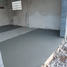 Wylewka betonowa w pomieszczeniu w trakcie realizacji, szalunek z desek widoczny przy krawędzi, gruz na zewnątrz, okno z luksferów w tle.