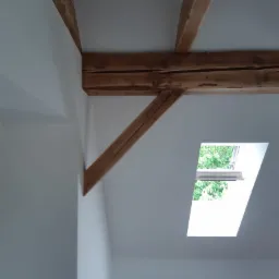 Wnętrze poddasza z drewnianymi belkami stropowymi, ścianami pomalowanymi na biało i oknem dachowym, przez które widać zieleń drzew.