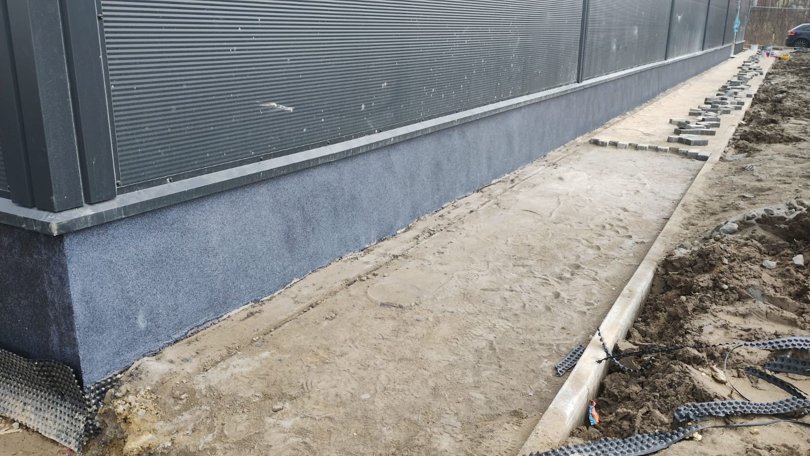 Szara elewacja budynku przemysłowego z widocznym fragmentem izolacji przy gruncie oraz przygotowanym podłożem pod chodnik z kostki.