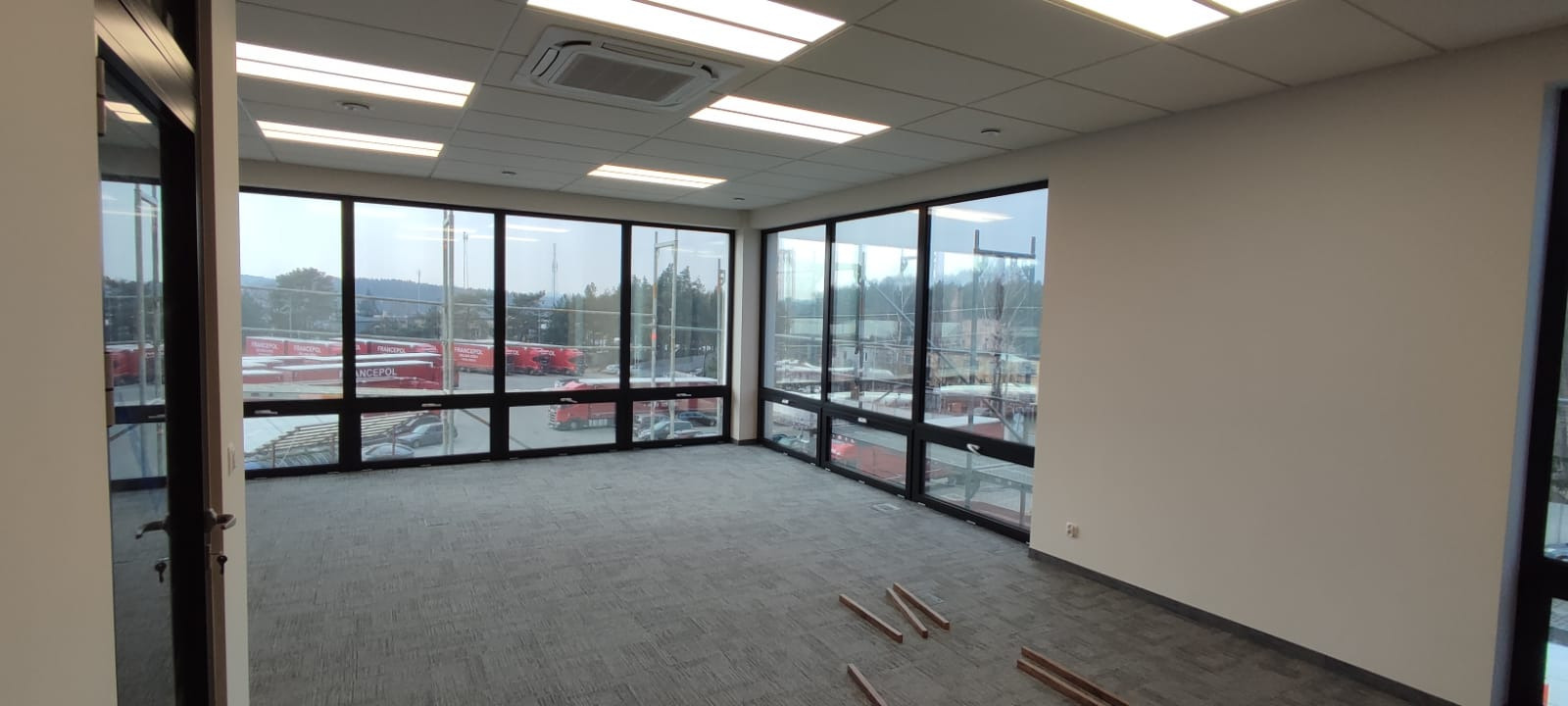 Wnętrze biurowe z dużymi oknami, widok na zewnątrz z czerwonymi ciężarówkami, szara wykładzina dywanowa i białe ściany, fragmenty listew przypodłogowych na podłodze.