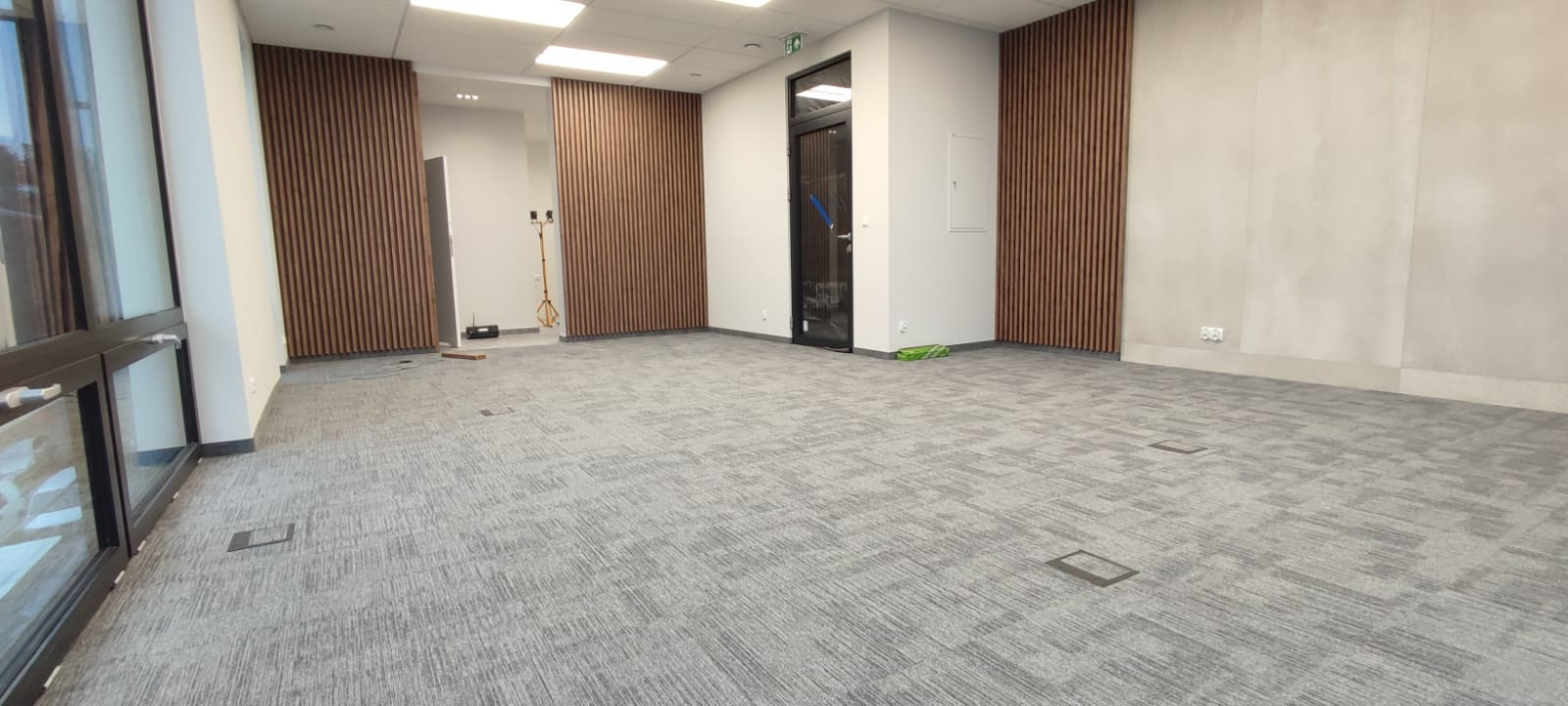 Wnętrze biurowe po remoncie: szara wykładzina dywanowa w geometryczny wzór, pionowe drewniane panele ścienne, przeszklone drzwi wejściowe i okna, jasne oświetlenie sufitowe.