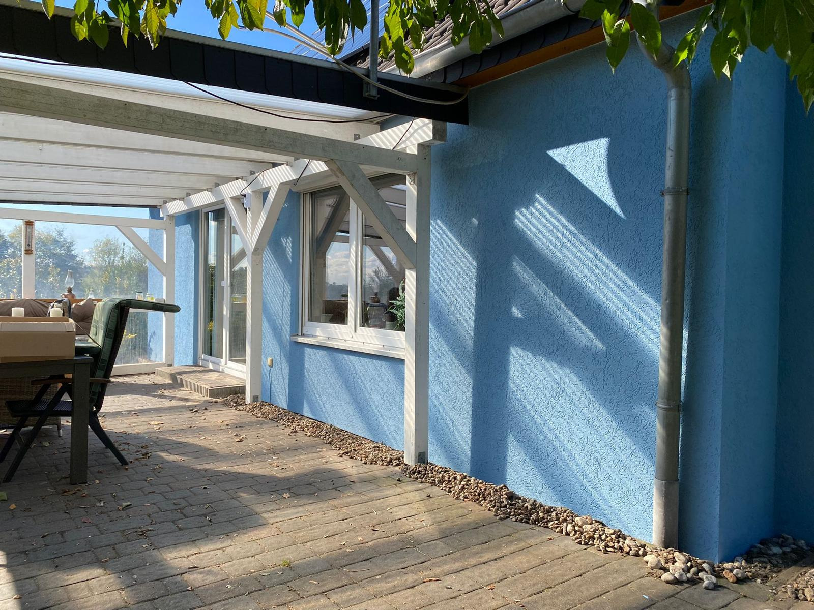 Błękitna, chropowata elewacja domu z oknem, widoczna z zewnątrz zadaszonego tarasu z kamienną podłogą, w słoneczny dzień.