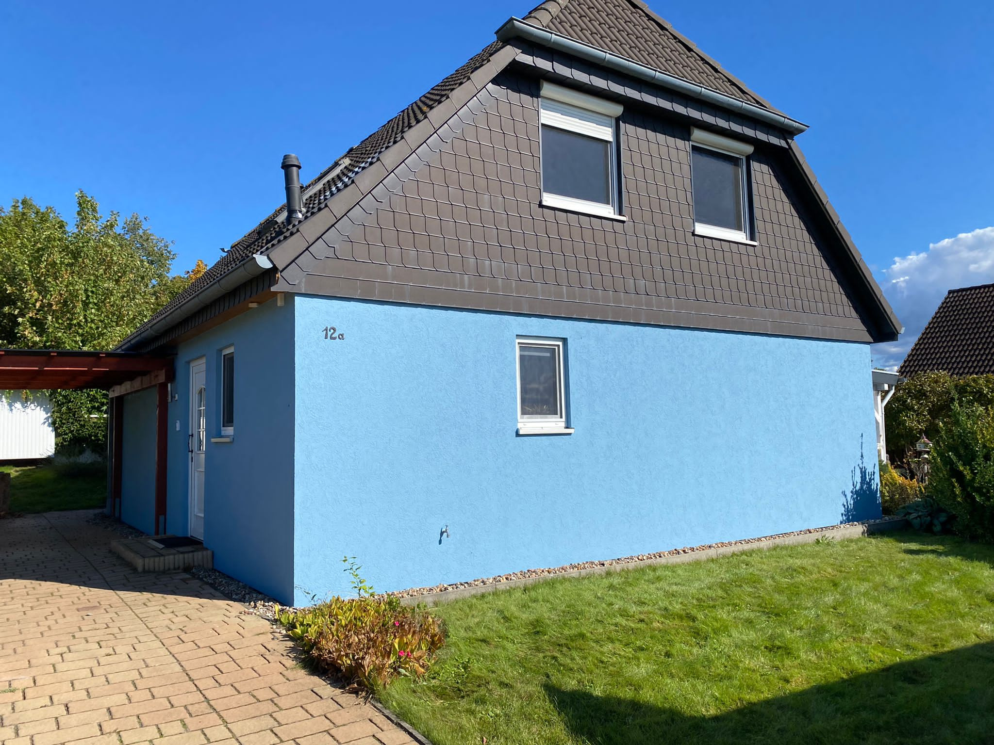Niebieski dom jednorodzinny z szarą dachówką karpiówką i numerem '12a' na elewacji, widok z trawnika w słoneczny dzień.