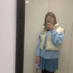 Osoba robi selfie w lustrze z psem na smyczy; pies ma czerwoną obrożę, a osoba ma na sobie niebieską bluzę, spódnicę i kremową kamizelkę puchową.