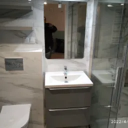 Wykończona łazienka z marmurowymi płytkami, umywalką w szarej szafce, toaletą i kabiną prysznicową; widoczne odbicie osoby robiącej zdjęcie w lustrze.