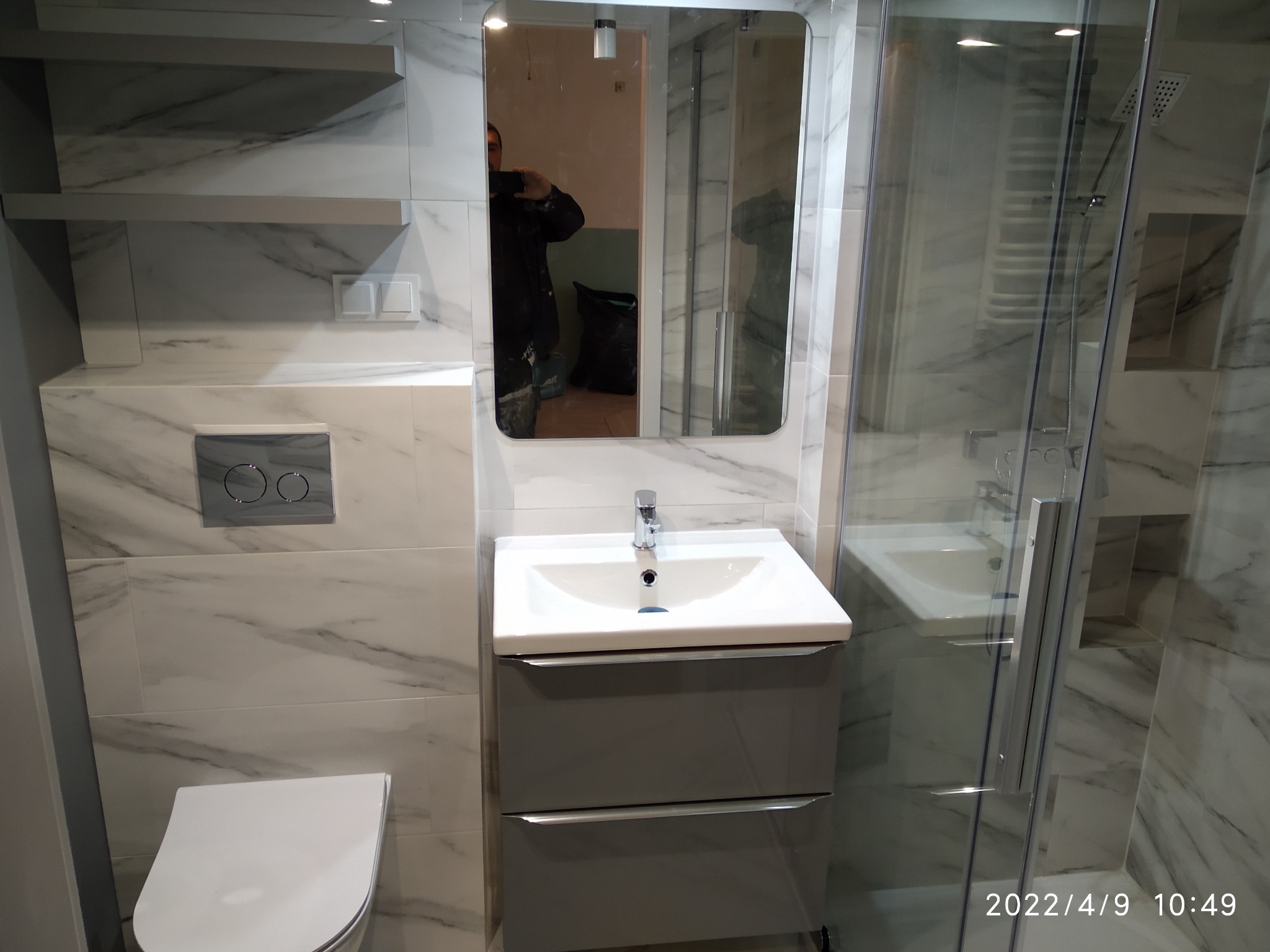 Wykończona łazienka z marmurowymi płytkami, umywalką w szarej szafce, toaletą i kabiną prysznicową; widoczne odbicie osoby robiącej zdjęcie w lustrze.