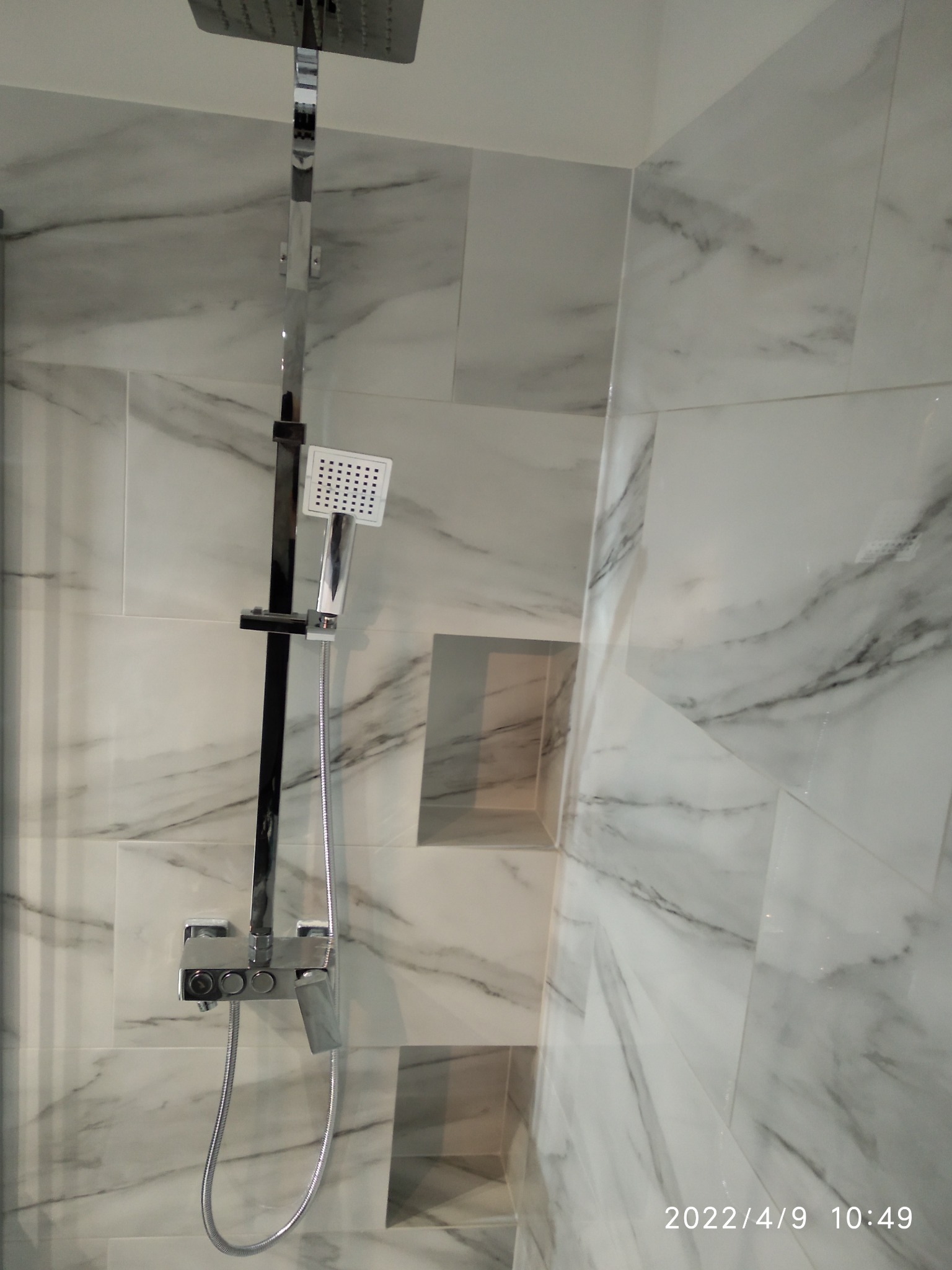 Bateria prysznicowa z deszczownicą i słuchawką na ścianie wyłożonej białymi płytkami imitującymi marmur z wnękami.