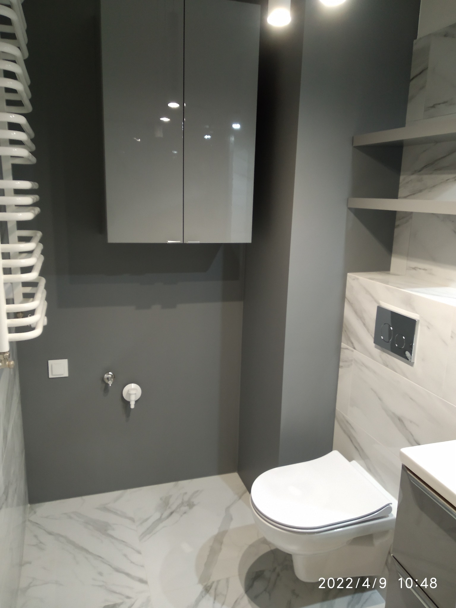 Nowoczesna łazienka w odcieniach szarości z marmurowymi płytkami, podwieszaną szafką i grzejnikiem drabinkowym, widok na toaletę wiszącą i minimalistyczną armaturę.