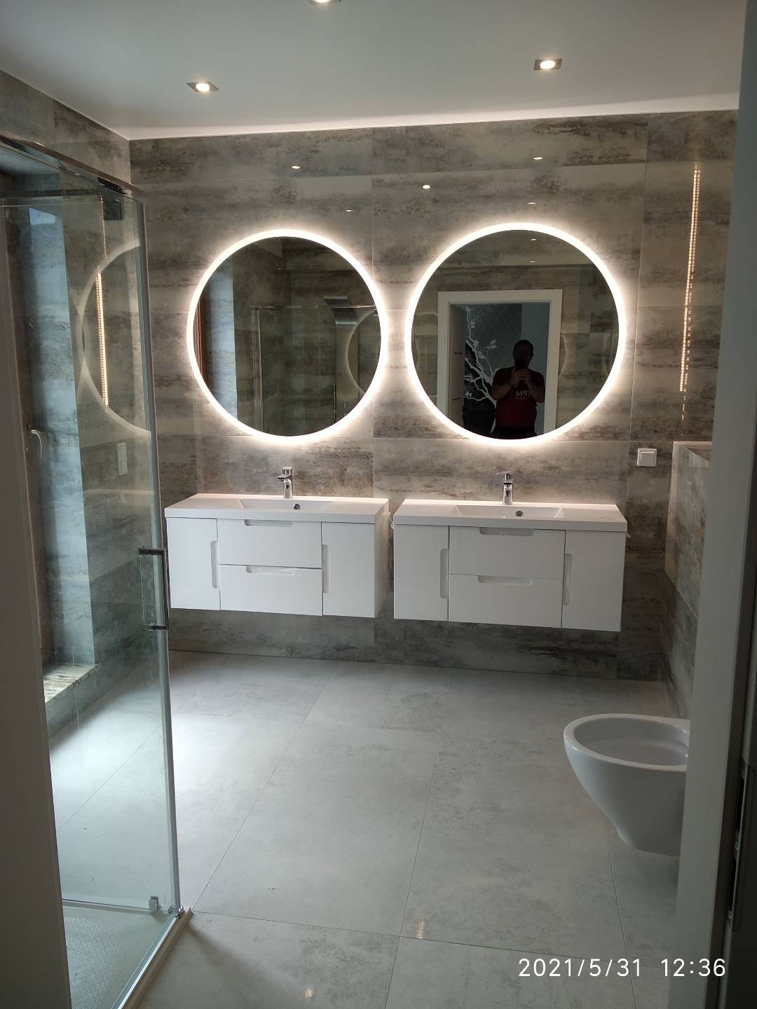 Nowoczesna łazienka z dwoma okrągłymi lustrami z podświetleniem LED, białymi szafkami pod umywalki i szarą glazurą imitującą kamień, kabina prysznicowa ze szkła hartowanego po lewej stronie.