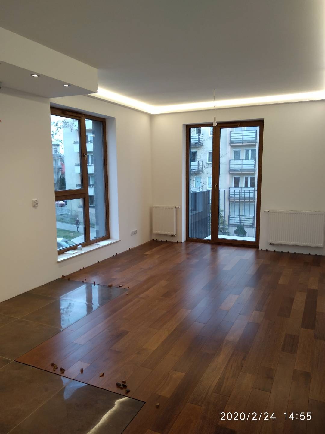 Wnętrze mieszkania w trakcie układania podłogi: panele drewniane w połowie ułożone, płytki ceramiczne w części pomieszczenia, widoczne kliny dystansowe, białe ściany i sufit z oświetleniem LED.