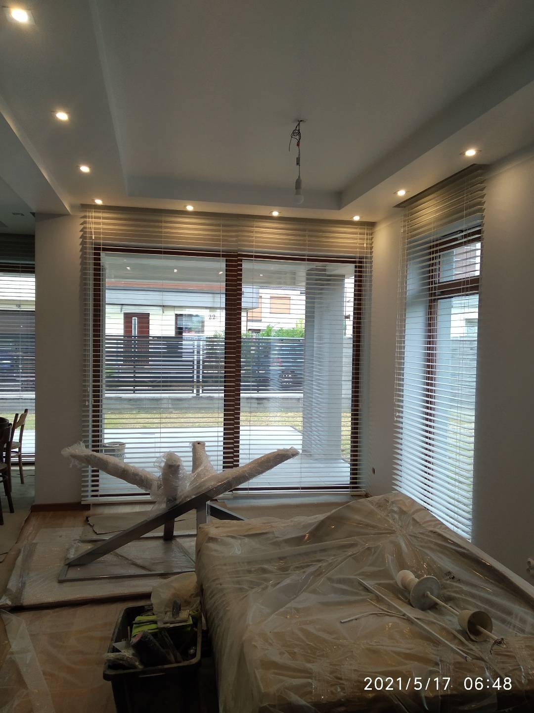 Pokój po montażu białych żaluzji poziomych w oknach z brązowymi ramami, meble zabezpieczone folią malarską, widoczne reflektory sufitowe i wisząca żarówka bez klosza.